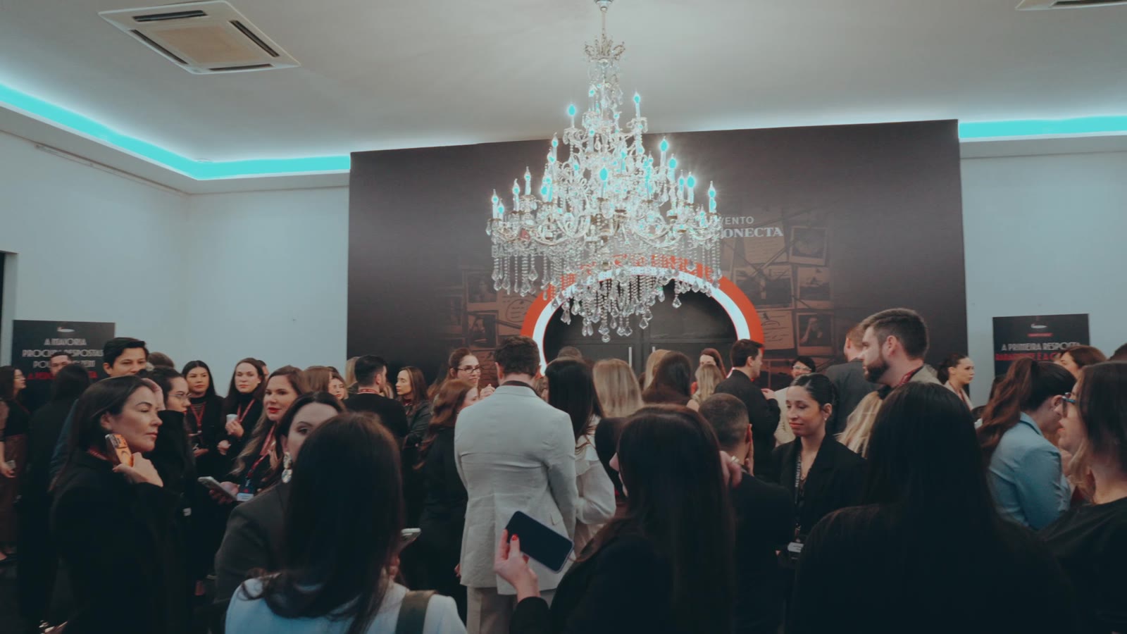 Sala do evento cheia, vista do fundo, com o lustre e o palco iluminado