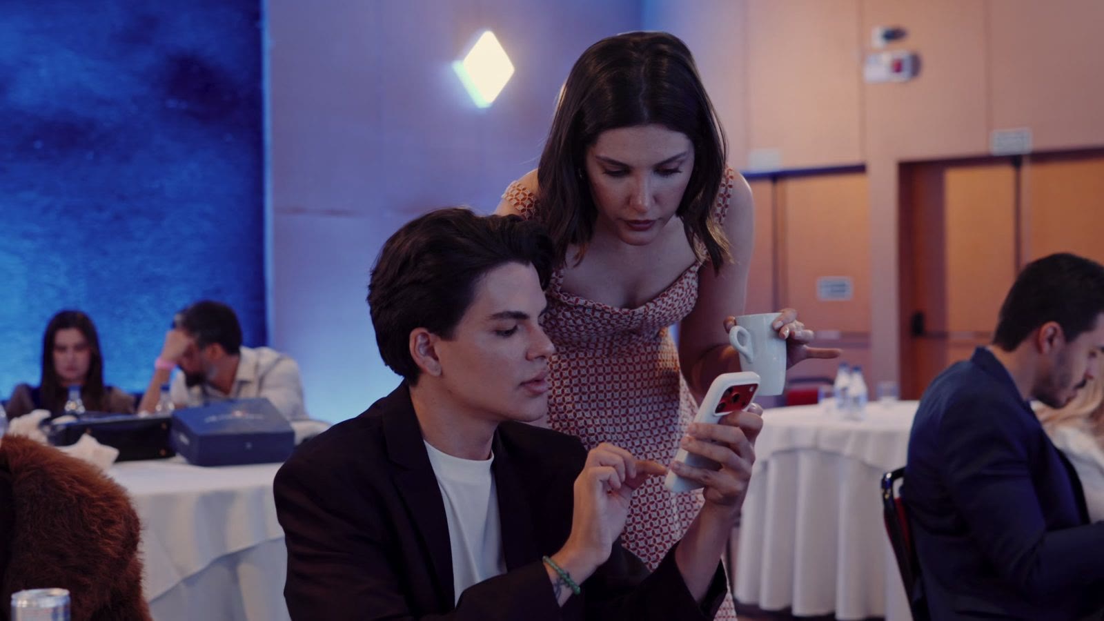 Ana Luiza em pé, ao lado de um participante sentado à mesa, olhando junto com ele a tela do celular