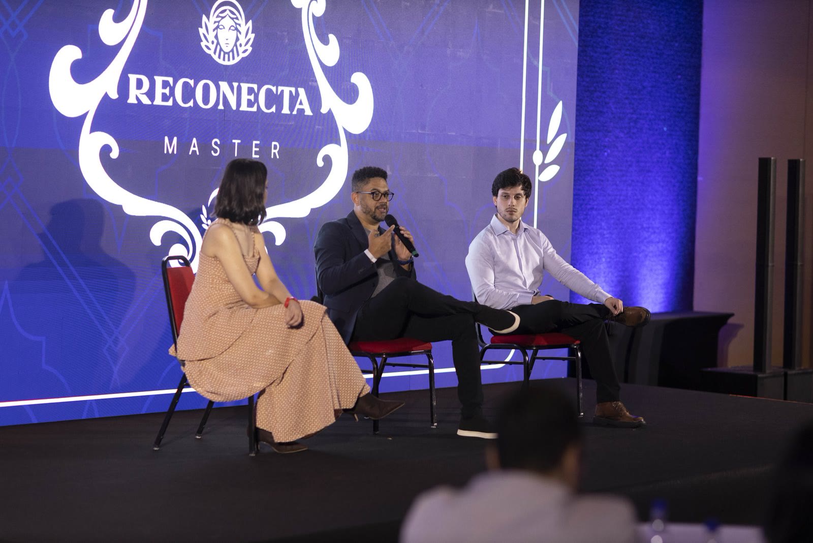 Palco do Reconecta Master, com o telão do encontro ao fundo e três pessoas sentadas conversando