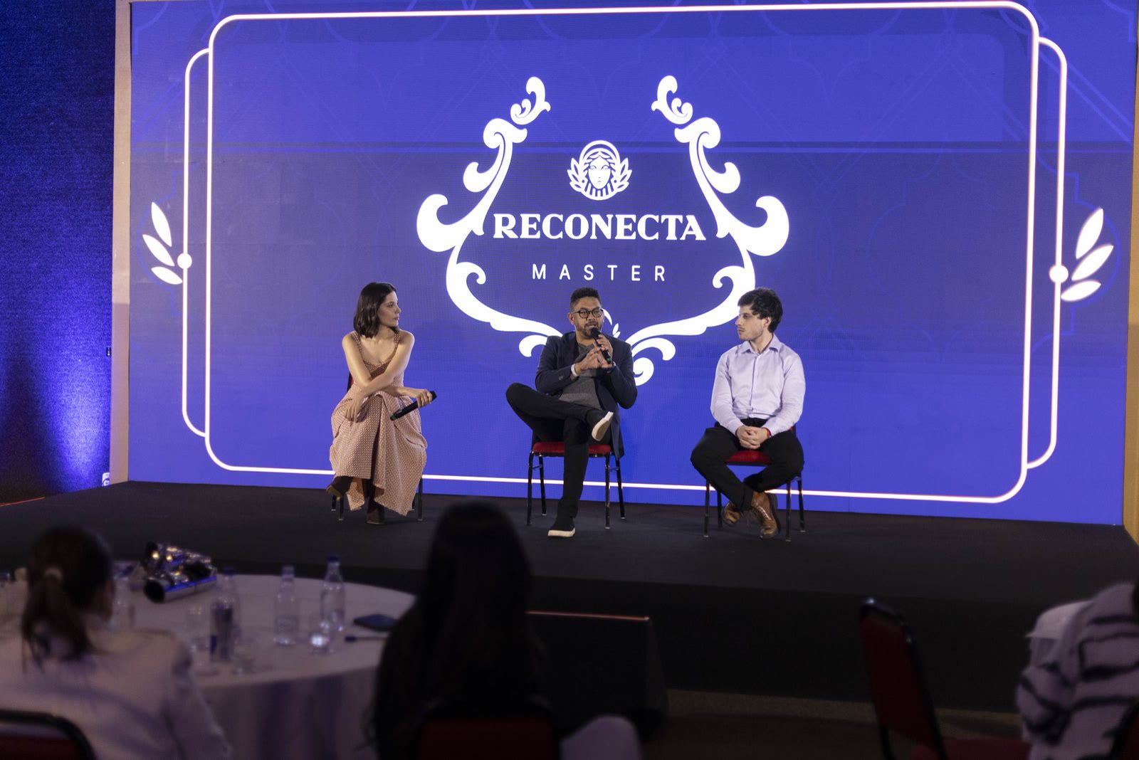 Palco do Reconecta Master: Ana Luiza, um convidado e Leonardo Rosso sentados diante do telão com o nome do encontro