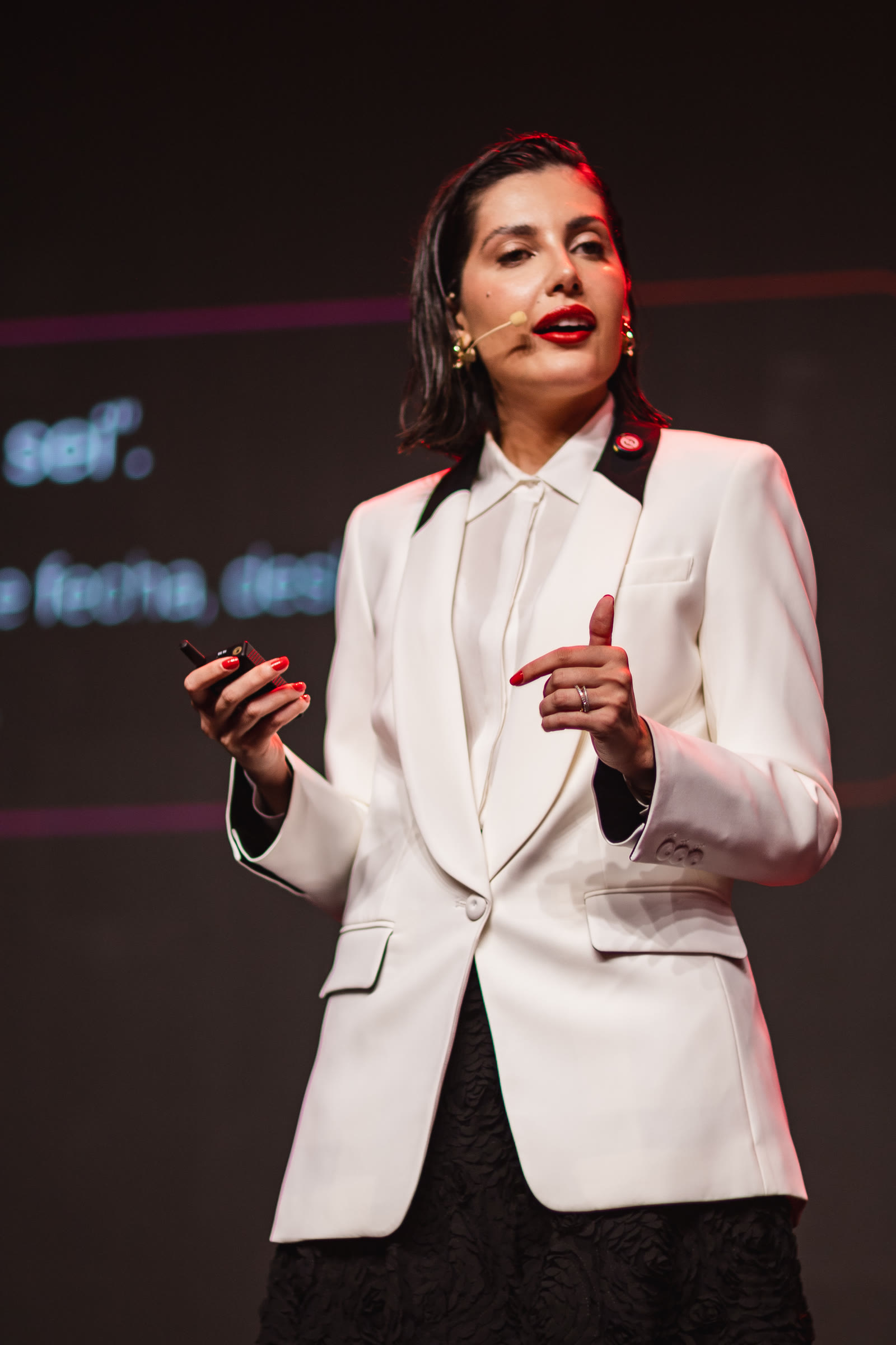 Ana Luiza falando no palco, de blazer branco, com o fundo vermelho do evento